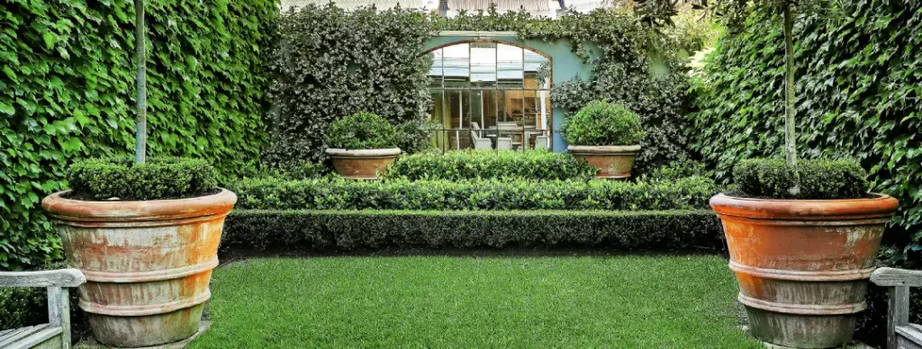 An arched garden mirror reflecting a patio area, framed by ivy walls and manicured boxwood hedges.